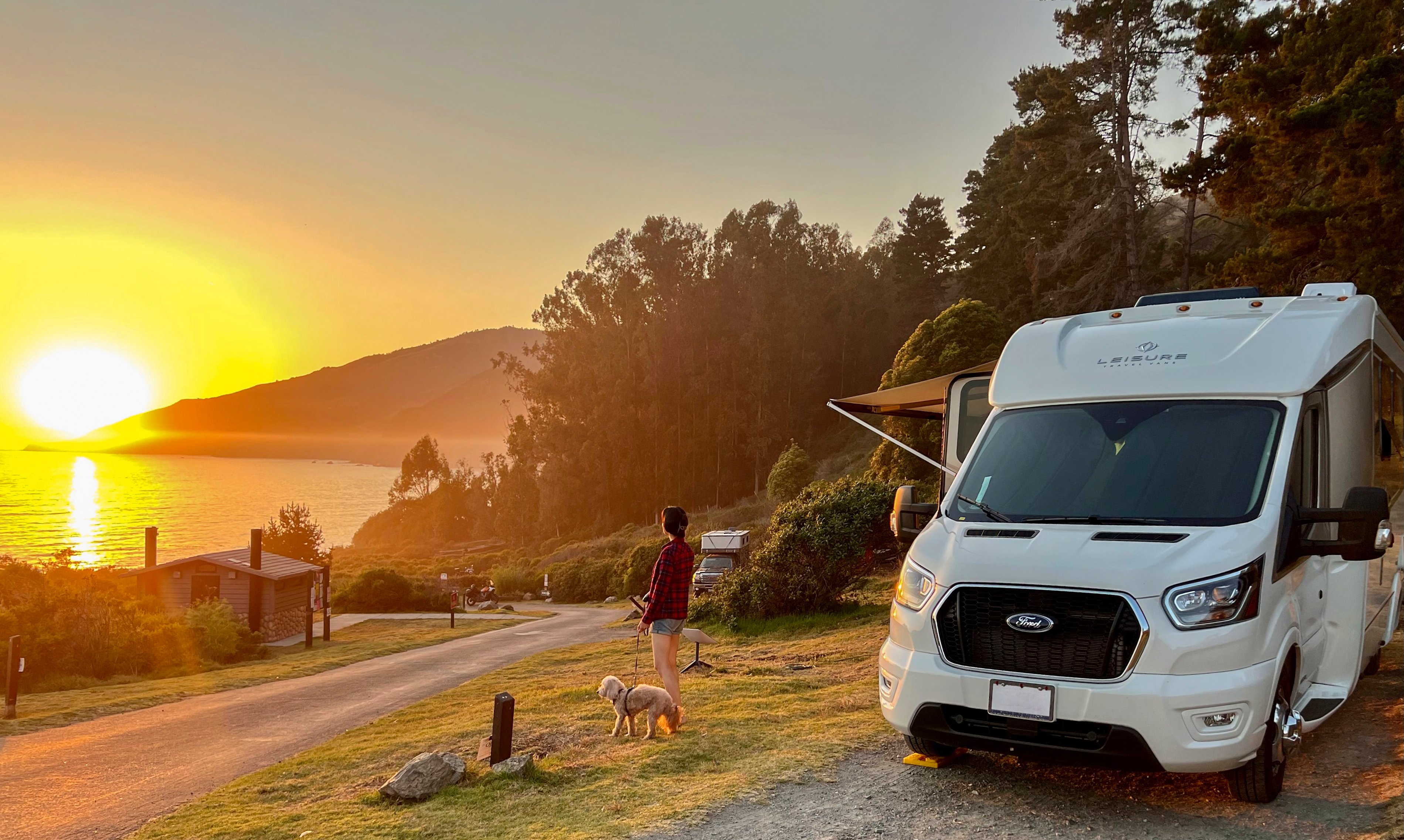 A woman and her dog stand beside their LTV and watch the sunset